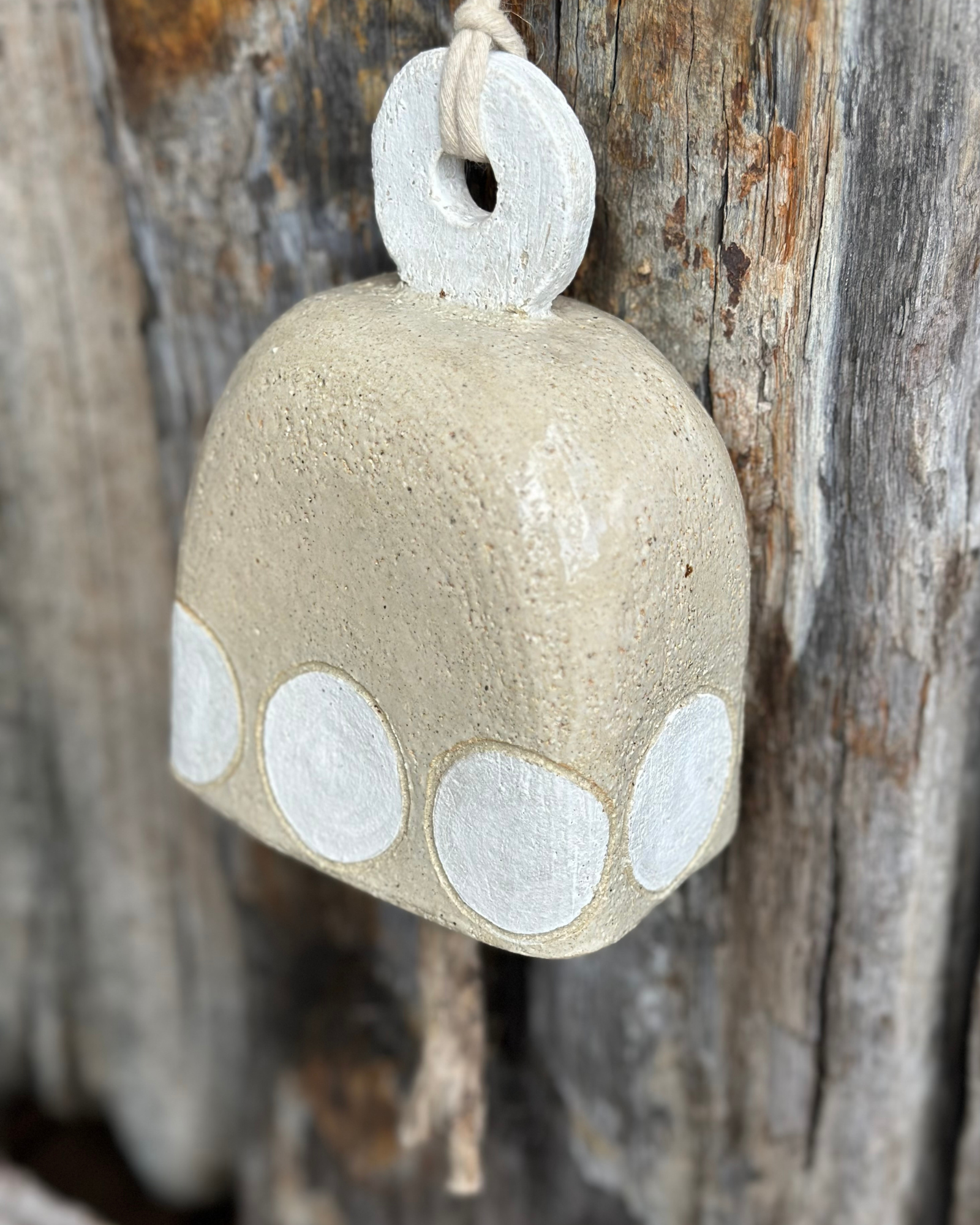 Handcrafted stoneware bell with white circular motif and driftwood striker, hanging against weathered timber backdrop