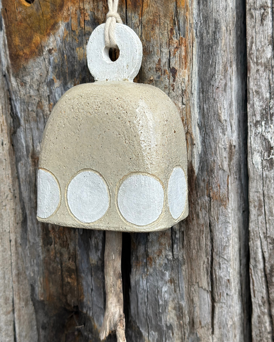 Handcrafted stoneware bell with white circular motif and driftwood striker, hanging against weathered timber backdrop
