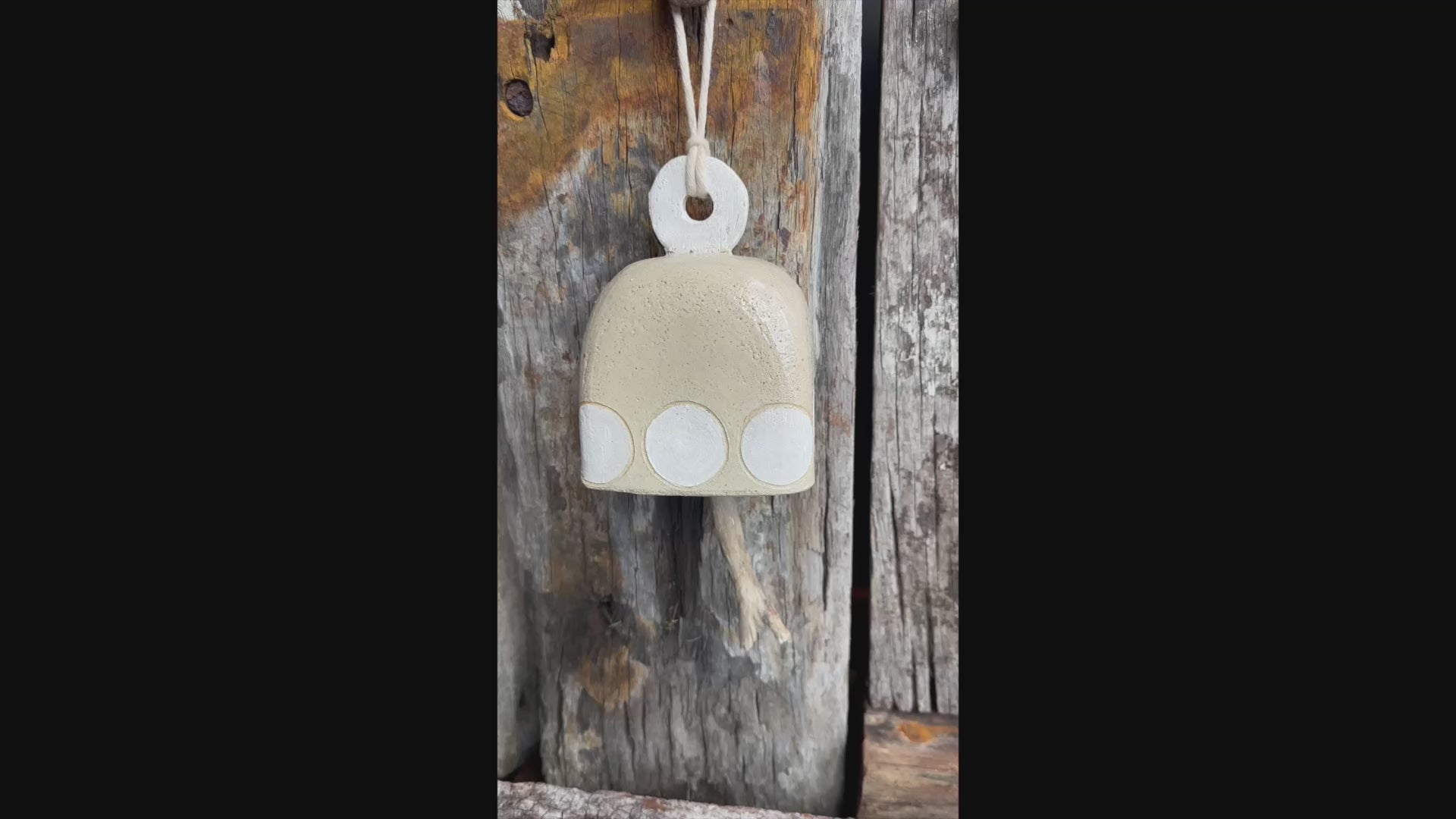 Sculptural ceramic bell in natural glaze with white circular detailing and driftwood striker, suspended on flax rope